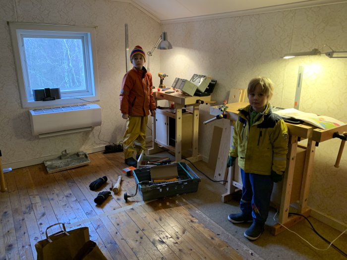 Två barn i ett arbetsrum med verktyg och en demonterad bordsåg på golvet.