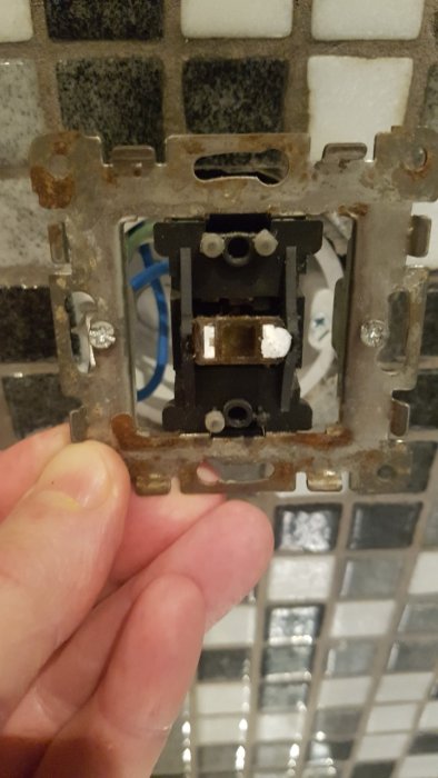 Hand håller en gammal och rostig ljusströmbrytare framför kakelvägg.