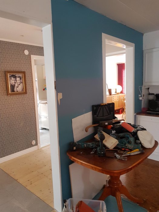 Husinteriör med en öppen dörröppning, väggar halvmålade i blått, verktyg på ett bord och en synlig passage till ett annat rum.