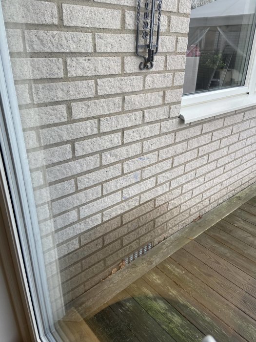 Fuktfläck på mexitegelvägg vid husrumshörn ovanför en träaltan, med en termometer fäst på väggen och ett fönster till höger.