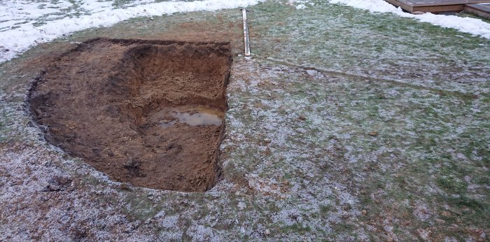 Grävd grop i trädgård för en framtida rund pool, snö på marken och en pöl av vatten synlig i gropen.