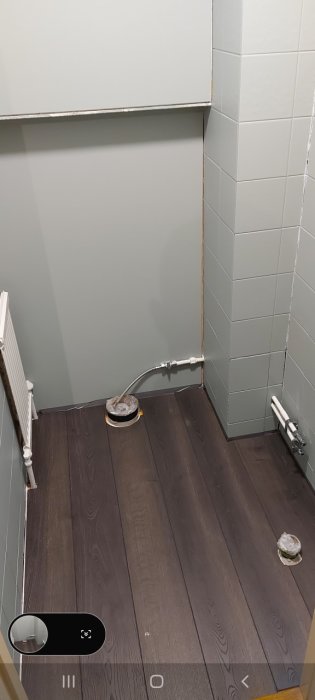 Hörn av ett badrum med synligt böjt rör, mörkt trägolv och gråvägg med vitt kakel.