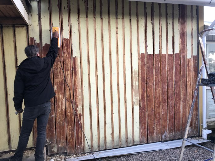 Person skrapar gammal färg på en träfasad under renovering, med delvis borttagna paneler och ställning.