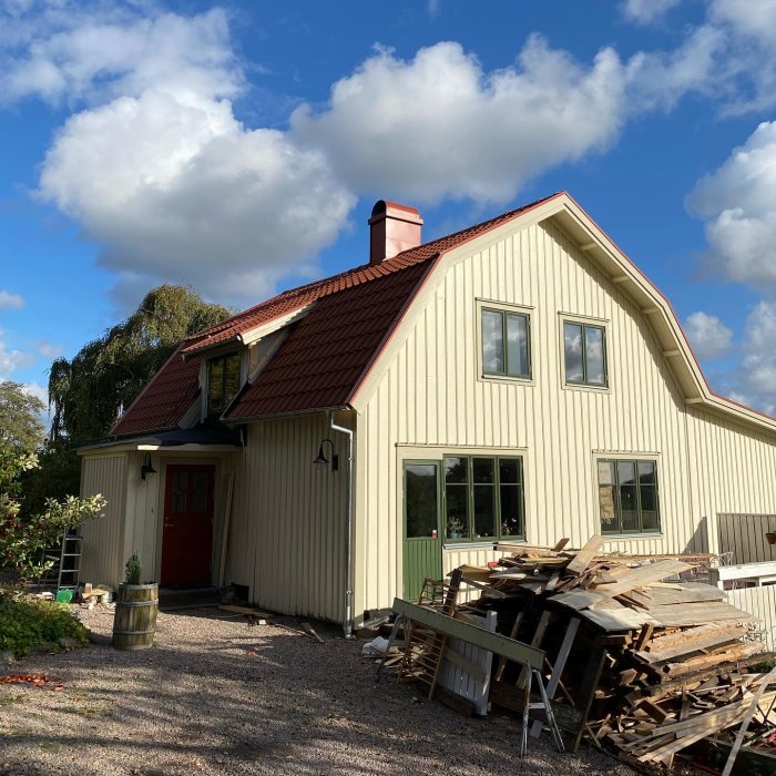 Ett nyrenoverat tvåvåningshus med ljus fasad och rött tak, en hög träskräp framför huset och blå himmel ovanför.