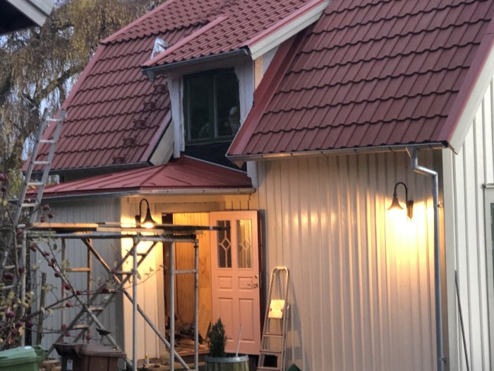 Renoverat hus från 1922 med rött tak och vit fasad i skymningen, ställning vid entrén.