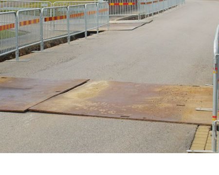 Metallplåtar som använts för tillfällig vägövergång över grävarbete på en gata, omgärdad av skyddsräcken.
