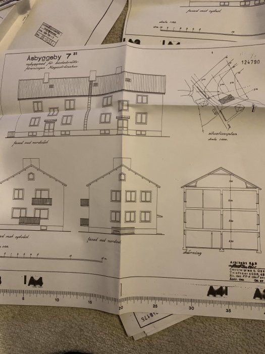 Arkitektritningar för en byggnad, inklusive fasadvyer och situationsplan, spridda på ett underlag med en linjal.