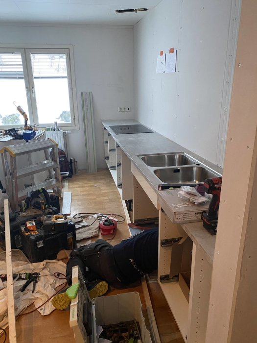 Köksrenovering pågår med verktyg, byggmaterial och en person som installerar skåp.