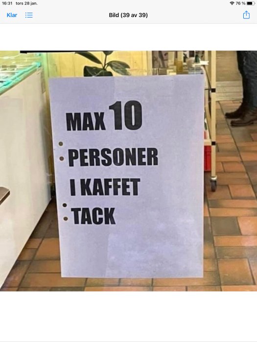 Skylt med texten "MAX 10 PERSONER I KAFFET TACK" inne i ett café.