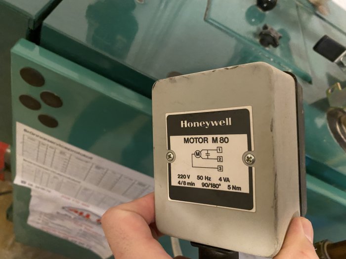 En Honeywell Motor M80 styrenhet hålls framför en grönmålad värmepanna, med tekniska specifikationer synliga.