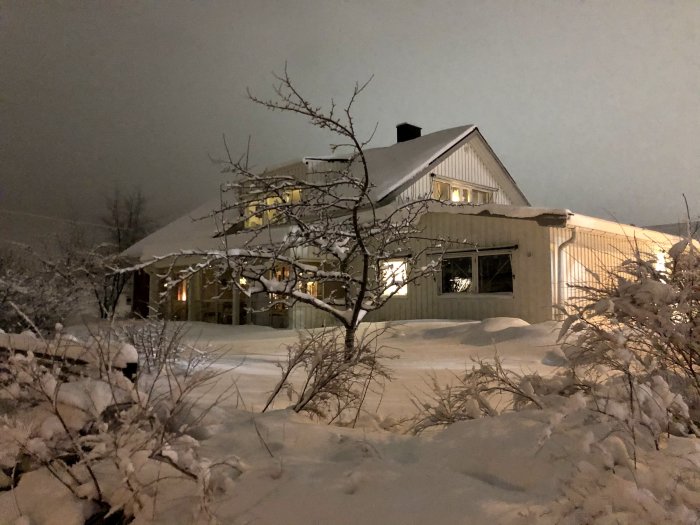 Ett hus täckt med snö på natten, väl upplyst inifrån, och omgivet av snöiga buskar.