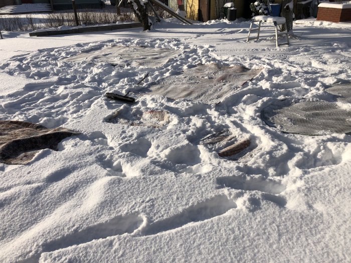 Mattor ligger i snön för rengöring i en trädgård med snötäckt mark och en vit trädgårdsstol i bakgrunden.