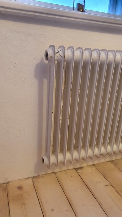 Gammal radiator med synliga anslutningsrör vid en vägg och trägolv.