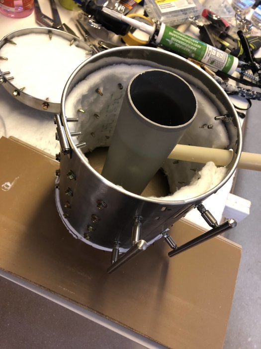 Rostfri pedalhink modifierad med beslag och skruvar, fiberisolering och ett centralt avloppsrör mot en verktygsrörig bakgrund.