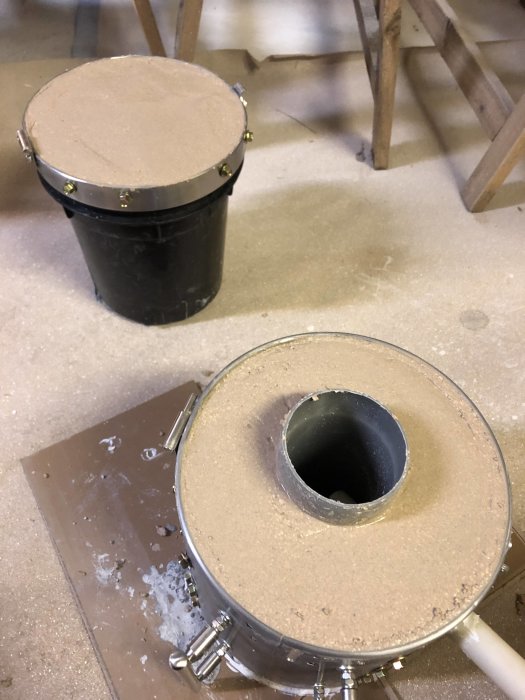 Rostfri pedalhink modifierad till isolering med plåtskruv, fiberisolering och lerkomposition, ett brännarrör bifogat vid sidan.