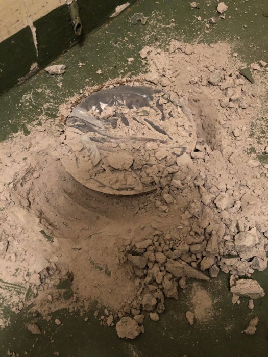 Golvbrunn omgiven av uppbruten betong och grus, med synlig rostig ring och armeringsjärn.