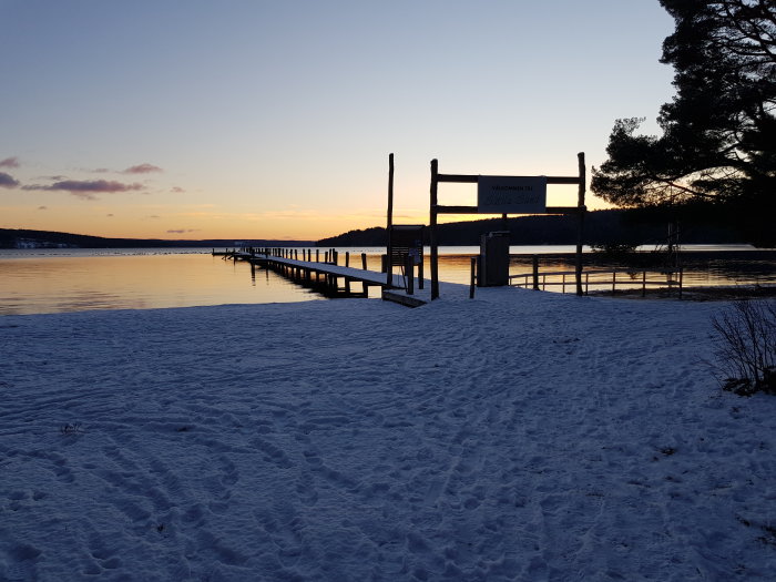 Solnedgång över en snötäckt strand med brygga och skylt som säger "Välkommen till Stilla Sand".