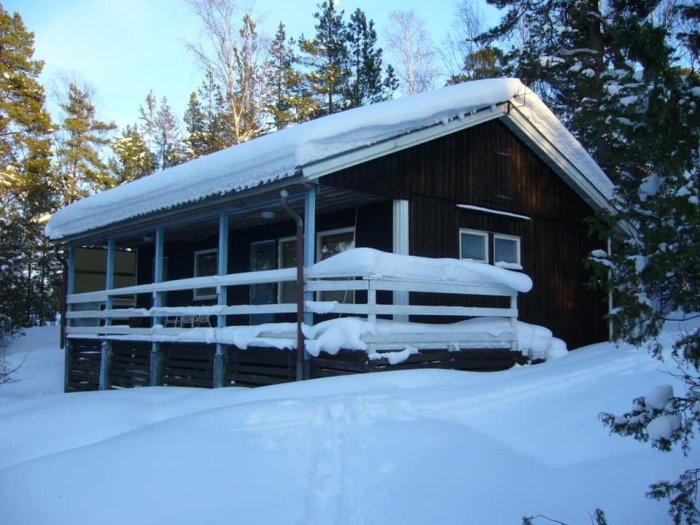 Trästuga med snötäckt tak och veranda omgiven av snöklädda träd och mark.