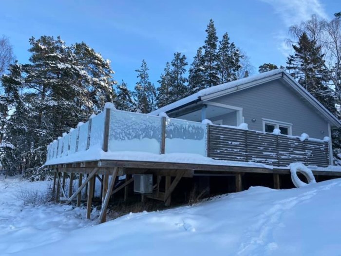 Vit stuga med veranda täckt av snö omringad av snöklädda träd och klarblå himmel.