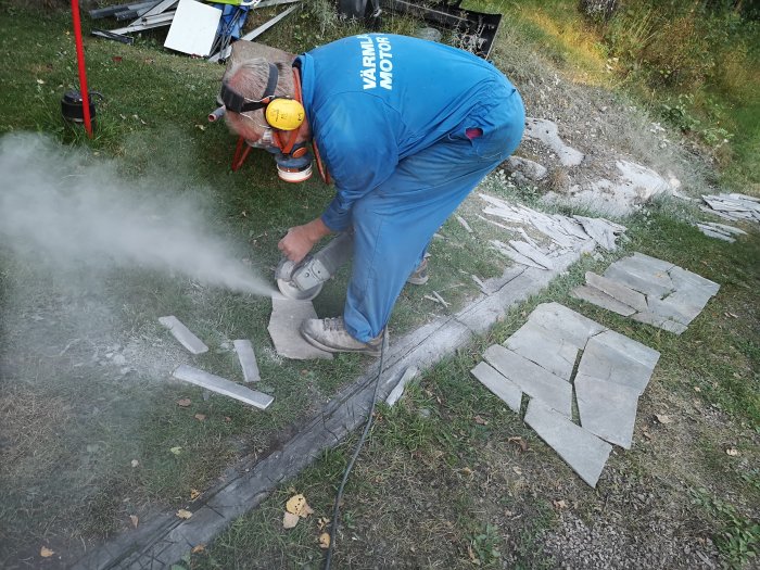 Person klipper takskiffer med vinkelslip, bär skyddsutrustning, skivor på marken.