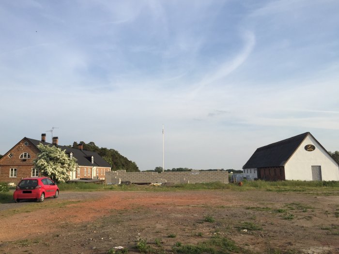 Röjt område med pågående byggnation av mur, röd bil i förgrunden, hus i bakgrunden under kvällshimmel.