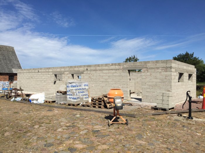 Hus under konstruktion med betongblock och murbruk, cementblandare i förgrunden och byggmaterial på marken.