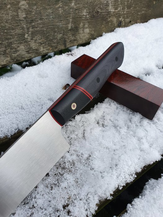 Handgjord 220 mm kockkniv i ståltypen 14c28n, med svart rödhandtag, vilande på träbit i snö.