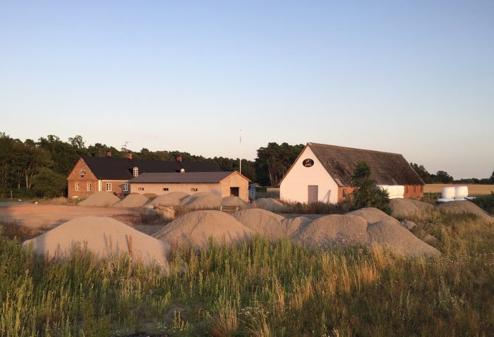 Lantgård med tegelhus och vit ladugård omgärdad av grushögar i gyllene kvällsljus, träd i bakgrunden och fält i förgrunden.