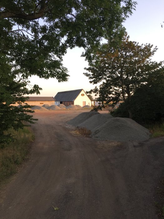 Gårdsbild med grushögar och väganläggning i förgrunden, byggprojekt med stort hus i bakgrunden under solnedgång.