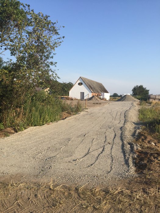 En nyanlagd grusväg som leder mot en vit lada med halmtak, omgiven av öppet fält och grön vegetation.