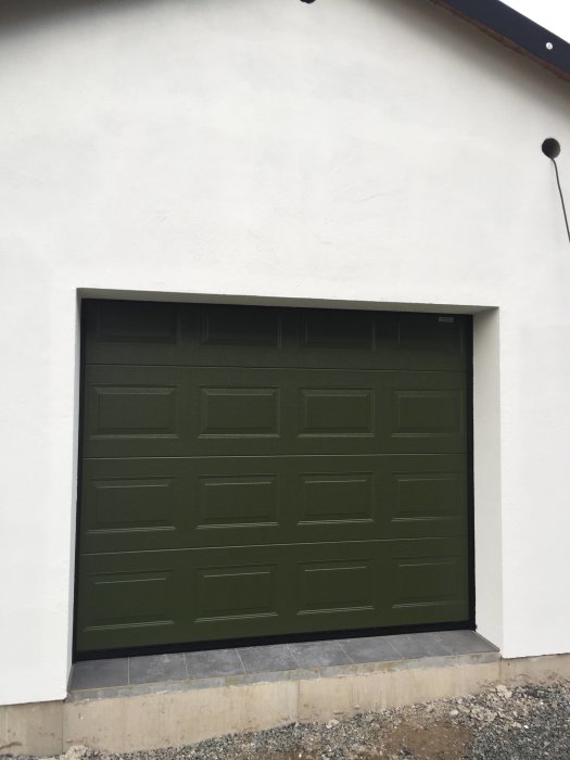 Nyinstallerad grön garageport i en vitmålad fasad på en verkstad med nylagt fall i klinker framför.