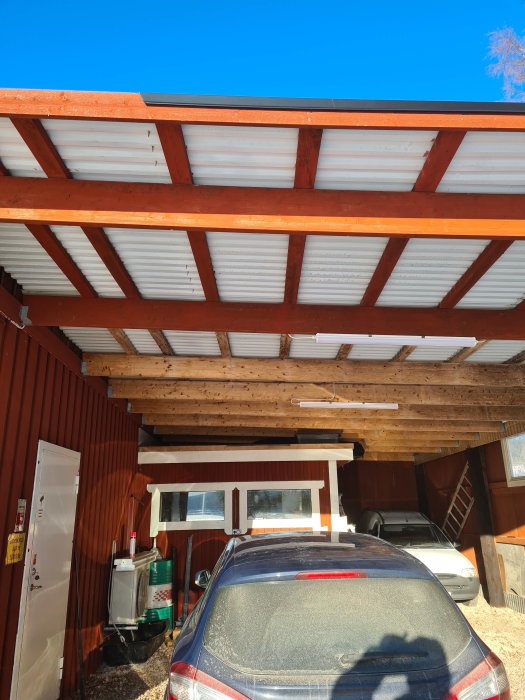 Utsikt under en carportstak med synliga takplåtar och träbjälkar, ovanför parkerade bilar.