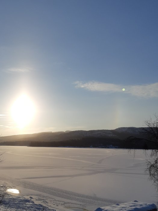 Vinterlandskap vid solnedgång med snötäckt sjö och skogklädda kullar i bakgrunden.