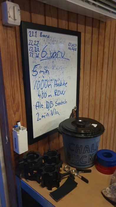 Whiteboard i gym med träningspass antecknat, viktskivor och kritburk i förgrunden.