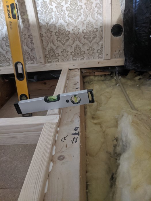 En påskruvad regel vid en deformerad bjälke i ett renoveringsprojekt, med en vattenpass som visar ojämnheten.