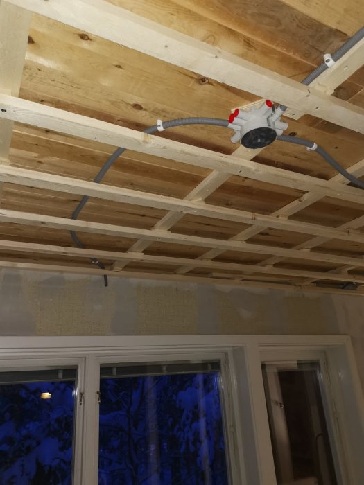 Installation av ljusfixture med dragna rör i ett oavslutat träbjälklag med synligt isoleringsmaterial ovanför ett fönster.