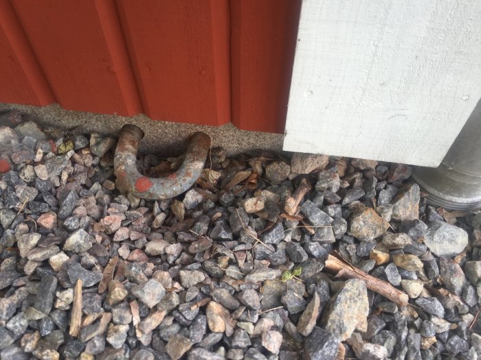 Metallögla för säkerhetslåsning inbäddad i sten och grus vid en husvägg.