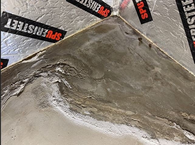 Ett hörn av ett betonggolv under renovering med tecken på fuktgenomträngning och skadat flytspackel längs väggarna.