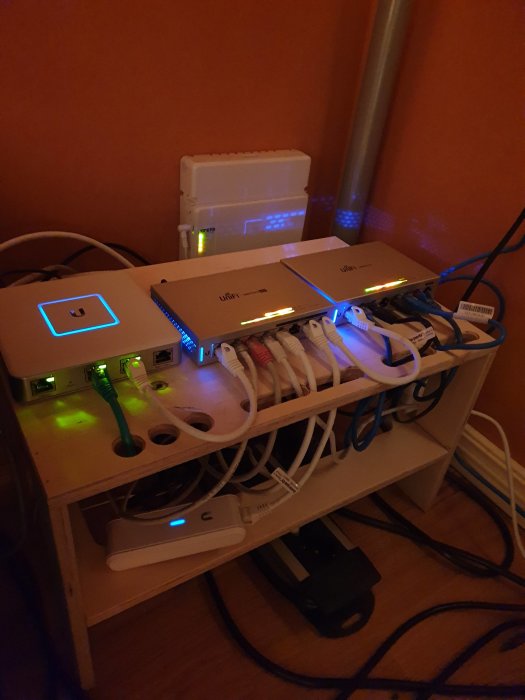 Nätverksuppställning under skrivbord med Unifi-utrustning och kablar på en hylla, indikerande en hemmanätverksinstallation.