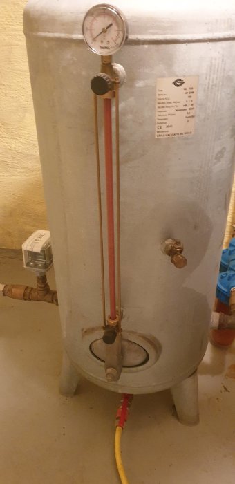 Grå hydrofortank med manometer och transparent rör på framsidan i en källare, synlig ventil på mitten.