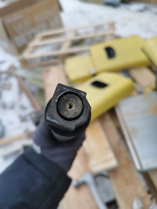 Hand håller en förstörd byggdetalj med töjt fästhål framför en bakgrund av byggmaterial.