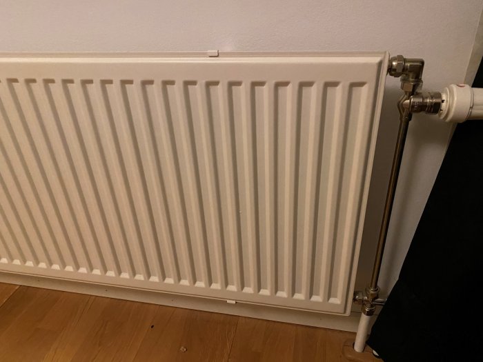 Vit radiator installerad vid vägg, kopplingsrör syns till höger, golv och vägg i bakgrund.