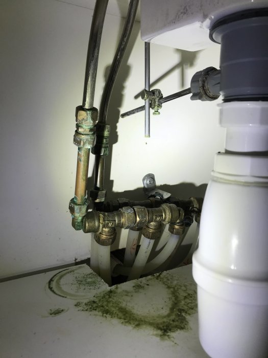 Vattenledningar och avloppsrör under handfat med synlig grönaktig missfärgning på skåpets botten.