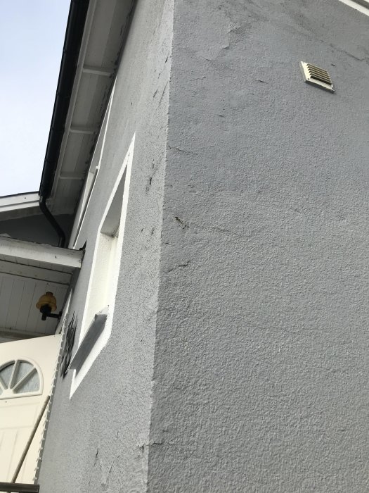 Vägg på hus med flagande fasadfärg och en ventilationsspjäll nära takfoten.