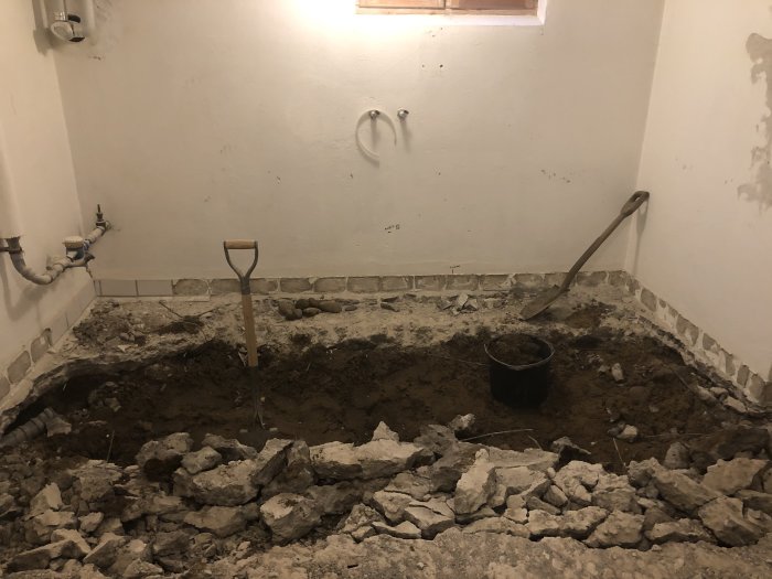 En delvis uppgrävd källargolv med synliga rör, en spade, och en hink bland grus och jord vid vita väggar.