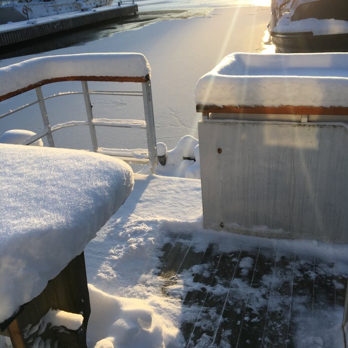 En snötäckt brygga med vit räcke och fotspår, vid en isbelagd flod i solljus.