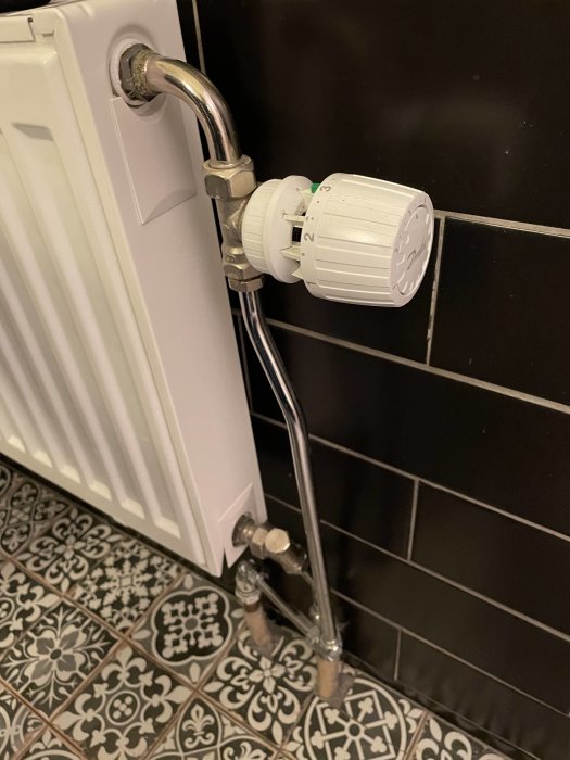 Termostatventil på en radiator monterad över ett kaklat golv med dekorativa mönster, intill en svart kaklad vägg.