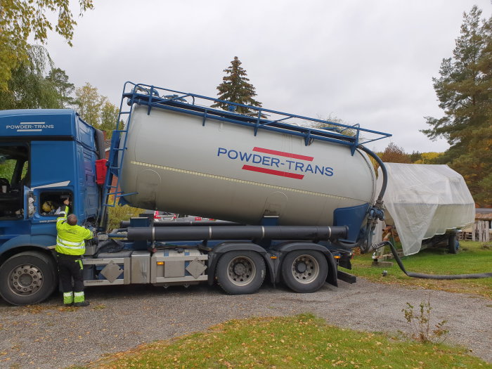 Lastbil med tank märkt "POWDER-TRANS" levererar material till ett skyddat område, med förare synlig.