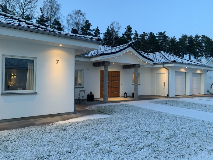 En vit modern villa med snöbeklädda takkanter och uppfart under vinterskymning.