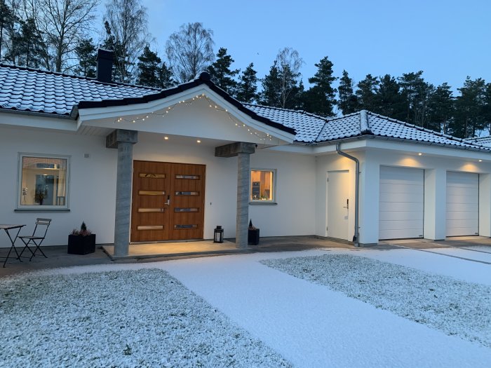 Ett hus täckt av snö vid skymning med upplyst entré och garage.